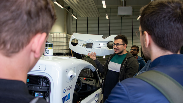 Ömer Dönmez shows a group something in the lab in front of an autonomous vehicle