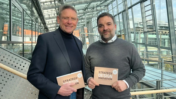Zwei Männer halten jeweils das Buch mit dem Titel „Strategy Design Innovation“, sie stehen in einem modernen gläsernen Gebäude.