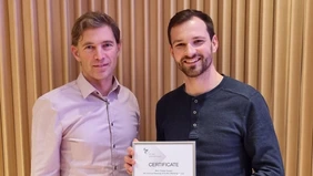 two men standing in front of an orange curtain, showing a framed certificate.