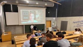 In a lecture hall, in the foreground the students from behind, in the background a man projected on a screen.