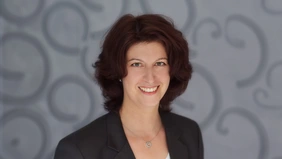 A woman with a black blazer in front of a gray background.