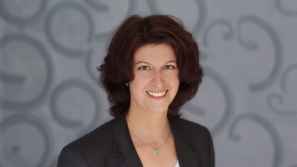 A woman with a black blazer in front of a gray background.